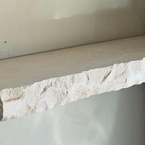 Op de afbeelding: Een rechthoekige, lichtgekleurde stenen plank met een ruwe, getextureerde rand. De plank is aan een muur bevestigd en toont zijn natuurlijke, ruwe uitstraling. De plank is ongeveer 91 cm lang.