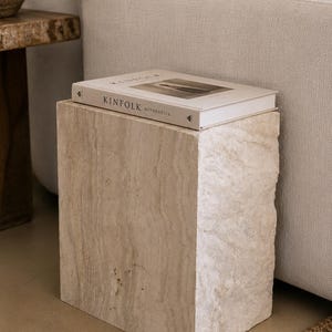 May include: A rectangular side table crafted from beige travertine stone with a natural, porous texture. Resting on top is a hardcover book titled Kinfolk Authentics, featuring a white cover with black text and a centered photographic image.