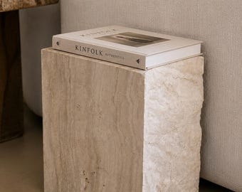 May include: A rectangular side table crafted from beige travertine stone with a natural, porous texture. Resting on top is a hardcover book titled Kinfolk Authentics, featuring a white cover with black text and a centered photographic image.