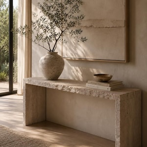 May include: A rectangular console table made of beige travertine stone with a rough, textured edge. On top sits a large, round, speckled beige vase containing a leafy green branch, a small wooden bowl, and two stacked books with white covers.