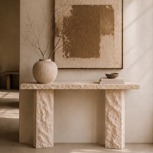 May include: A beige stone console table with a rough, chiseled texture on the top and two rectangular legs. On the table sits a round, ribbed ceramic vase containing thin, bare branches, alongside a small bowl resting on two stacked books.