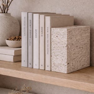 May include: A collection of books with neutral-toned covers, arranged on a wooden shelf. A textured, rectangular bookend in a light beige color supports the books. The titles are visible on the spines.