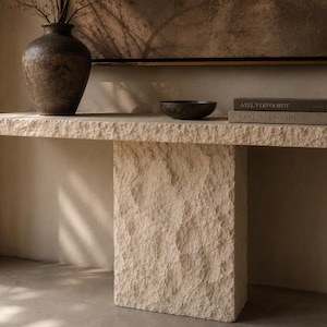 May include: A beige stone console table with a rough, chiseled texture and a central pedestal base. On the surface sits a dark brown ceramic vase, a small dark bowl, and two stacked books titled Axel Vervoordt and Aabl-Servoore.