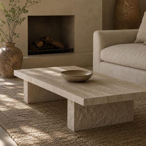 May include: A rectangular beige travertine coffee table with a smooth top surface and thick, textured slab legs. A shallow, speckled beige bowl sits in the center of the table. The stone features natural horizontal veining and a porous finish.