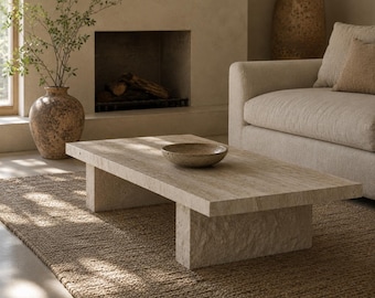 May include: A rectangular beige travertine coffee table with a smooth top surface and thick, textured slab legs. A shallow, speckled beige bowl sits in the center of the table. The stone features natural horizontal veining and a porous finish.