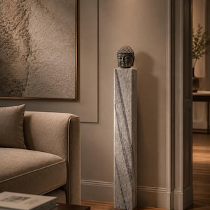May include: A dark gray stone Buddha head sculpture resting atop a tall, rectangular, light gray marble pedestal with diagonal veining. The sculpture features a textured, rounded top and serene facial features, standing as a singular unit.