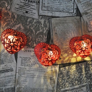 Puede incluir: Una cadena de luces rojas en forma de corazón con un diseño de encaje, colgando sobre un fondo de páginas antiguas impresas.