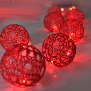 Puede incluir: Una cadena de luces de globo rojo de ganchillo con bombillas LED blanco cálido. Las luces están unidas por un cable transparente.