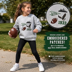 Könnte beinhalten: Ein hellgraues Langarm-Sweatshirt mit einem aufgedruckten Grafikdesign, das einen grünen Wimpel mit dem Text Philly, eine schwarze Stadtsilhouette, einen braunen Football, einen Adler und die Zahl 1933 zeigt, optisch wie Stickaufnäher.