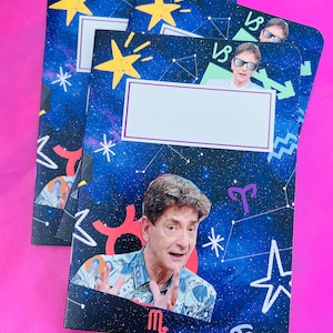 K&ouml;nnte beinhalten: Notizb&uuml;cher mit kosmischem Design, gelben Sternen, Sternbildern und Bildern eines Mannes. Die Notizb&uuml;cher haben ein wei&szlig;es rechteckiges Etikett zur Personalisierung. Der Hintergrund ist tiefblau mit wei&szlig;en Sternen und astrologischen Symbolen.