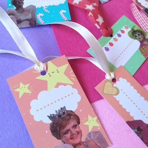 Può includere: Una collezione di etichette regalo colorate con vari disegni e immagini. Alcune etichette presentano una donna sorridente, stelle e bastoncini di zucchero. Le etichette sono attaccate a nastri e disposte su uno sfondo rosa, viola e bianco.