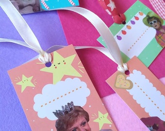 Könnte beinhalten: Eine Sammlung farbenfroher Geschenkanhänger mit verschiedenen Designs und Bildern. Einige Anhänger zeigen eine lächelnde Frau, Sterne und Zuckerstangen. Die Anhänger sind an Bändern befestigt und auf einem rosa, lila und weißen Hintergrund angeordnet.