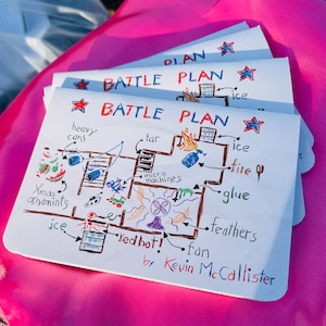 Puede incluir: Una pila de hojas de papel blanco con un diseño de "BATTLE PLAN" dibujado a mano. El plan incluye ilustraciones etiquetadas de "heavy cans", "tar", "fire" y "Xmas ornaments". El nombre del artista, Kevin McCallister, está en la parte inferior.