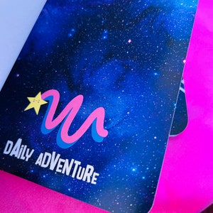 K&ouml;nnte beinhalten: Ein Notizbuch mit einem Design eines blauen Sternenhimmels. Das Cover zeigt ein stilisiertes "M"-Logo in Pink und Blau, einen gelben Stern und die Worte "DAILY ADVENTURE" in Wei&szlig;. Das Notizbuch befindet sich vor einem hellrosa Hintergrund.