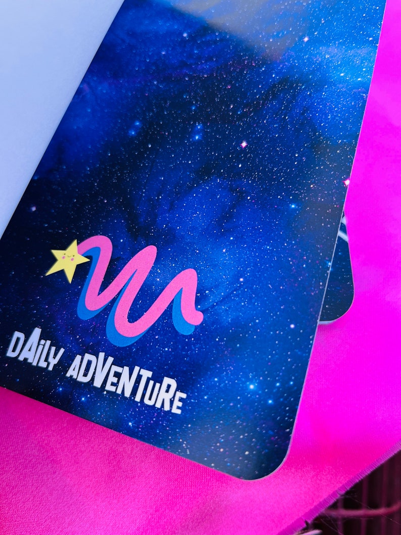 K&ouml;nnte beinhalten: Ein Notizbuch mit einem Design eines blauen Sternenhimmels. Das Cover zeigt ein stilisiertes "M"-Logo in Pink und Blau, einen gelben Stern und die Worte "DAILY ADVENTURE" in Wei&szlig;. Das Notizbuch befindet sich vor einem hellrosa Hintergrund.