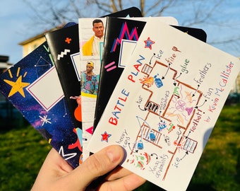 Könnte beinhalten: Eine Hand hält einen Stapel Notizbücher mit verschiedenen Einbänden. Designs umfassen eine Galaxie, geometrische Formen und Porträts. Ein Notizbuch zeigt "BATTLE PLAN" und eine handgezeichnete Skizze mit Begriffen wie "tar" und "ice".