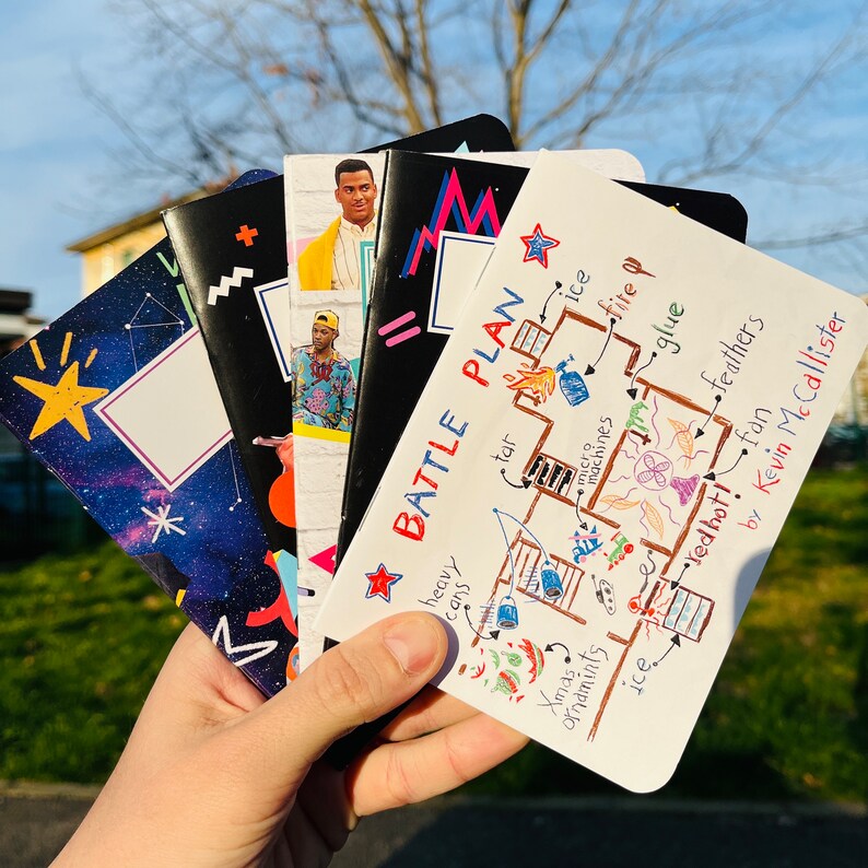 K&ouml;nnte beinhalten: Eine Hand h&auml;lt einen Stapel Notizb&uuml;cher mit verschiedenen Einb&auml;nden. Designs umfassen eine Galaxie, geometrische Formen und Portr&auml;ts. Ein Notizbuch zeigt "BATTLE PLAN" und eine handgezeichnete Skizze mit Begriffen wie "tar" und "ice".