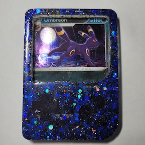 May include: A blue and black Pokemon card featuring Umbreon, a dark-type Pokemon, with the text "Umbreon" and "Evolves from Eevee". The card is encased in a blue resin frame with glitter.