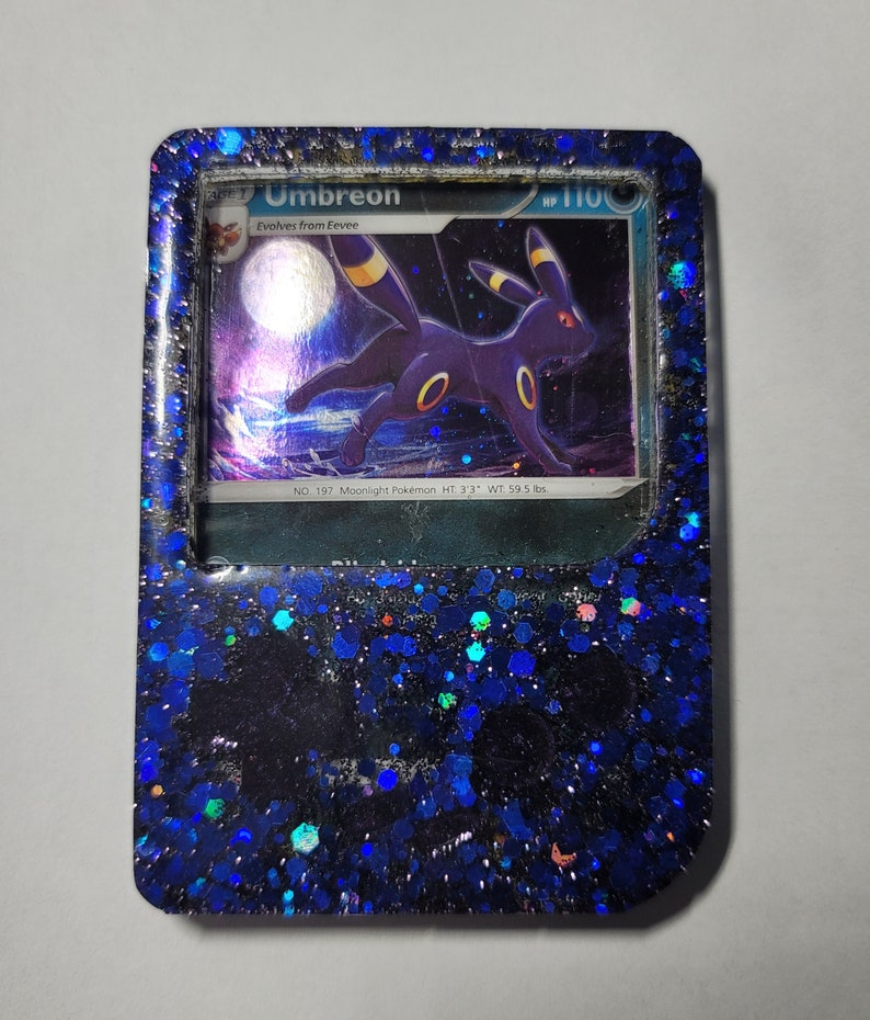 May include: A blue and black Pokemon card featuring Umbreon, a dark-type Pokemon, with the text "Umbreon" and "Evolves from Eevee". The card is encased in a blue resin frame with glitter.