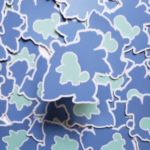 May include: A pile of blue and light green stickers featuring a cartoon character. The character is a blue creature with a light green head and body. The stickers are all the same design and are stacked on top of each other.