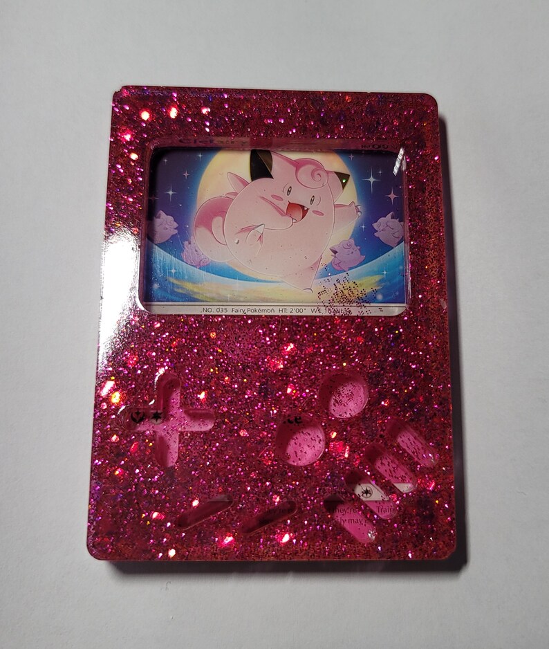 May include: Pink glitter resin Game Boy with a clear frame and a Jigglypuff Pokemon card inside. The card shows Jigglypuff floating in the air with a moon and stars in the background. The text on the card reads "NO. 035 Fairy Pokemon HT: 2'00" WK: 19.8 lbs."