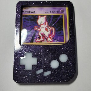 May include: A black and purple Game Boy Color-style case with a clear window displaying a Pokemon card featuring Mewtwo. The case has white buttons and a sparkly finish.
