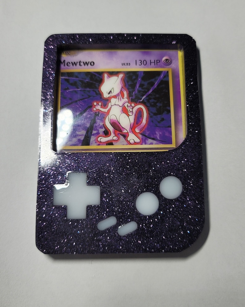 May include: A black and purple Game Boy Color-style case with a clear window displaying a Pokemon card featuring Mewtwo. The case has white buttons and a sparkly finish.