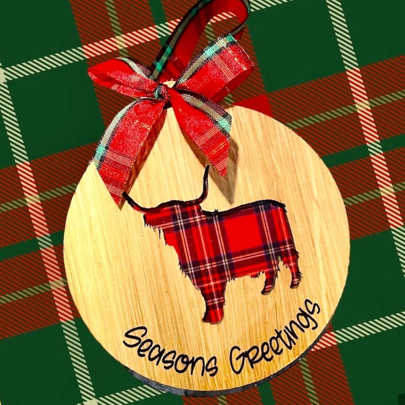 May include: A wooden Christmas ornament featuring a red and black plaid Highland cow with the text "Seasons Greetings" on the bottom.