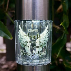 Könnte beinhalten: Solarbetriebene Gedenkleuchte mit gebürstetem Metallaufsatz und transparentem Glaskörper. Das Glas ist mit den Worten "I Have Angels in Heaven, I Call Them Mum & Dad" und Engelsflügeln graviert. Grünes Laub im Hintergrund.