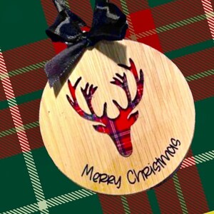 May include: A round wooden ornament with a red and black plaid reindeer cutout and the text "Merry Christmas" written in black.