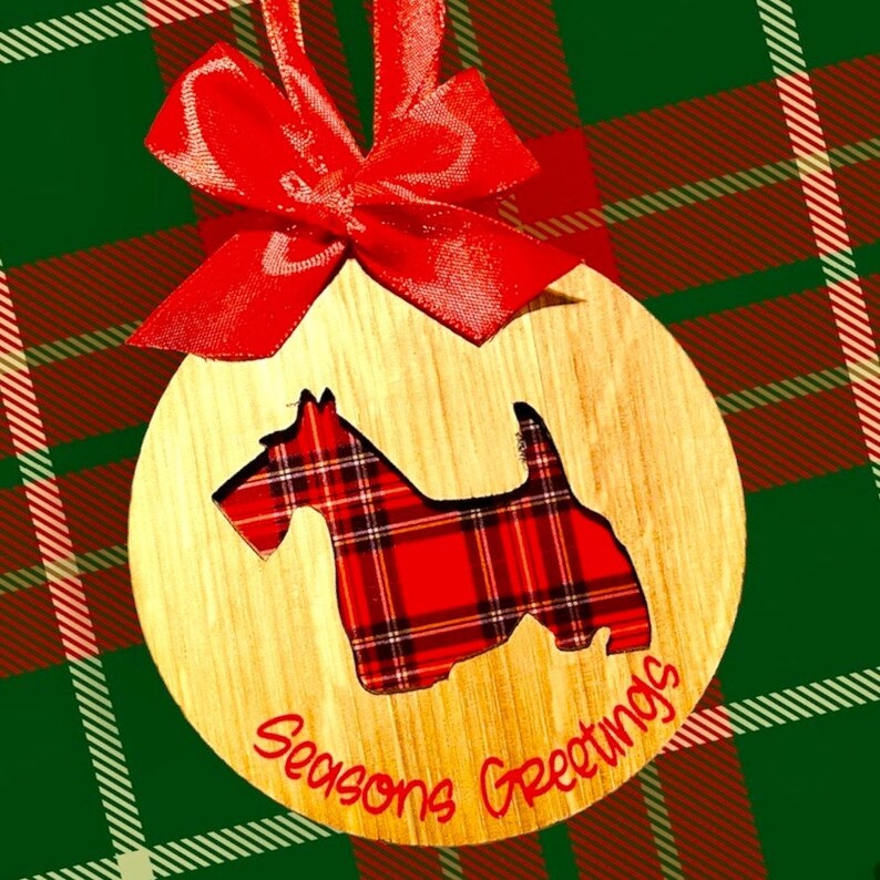 May include: A wooden Christmas ornament with a red and black plaid dog silhouette and the text "Seasons Greetings". The ornament is decorated with a red bow.