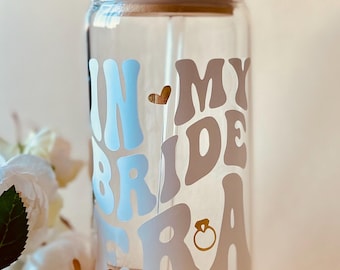 Könnte beinhalten: Ein durchsichtiger Glasbecher mit einem Holzdeckel und einem Strohhalm. Der Becher hat einen weißen Vinyl-Aufkleber mit der Aufschrift "In My Bride Era" mit einem kleinen Herz und einer goldenen Ringgrafik.