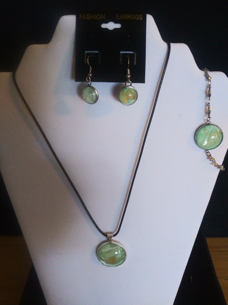 Op de afbeelding: Een groene en gouden set met ketting, oorbellen en armband met een zwarte koordsluiting en een zilveren kettingarmband. De sieraden hebben ronde hangers met een groen en gouden marmerpatroon.