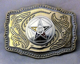 May include: A gold-toned belt buckle with a silver five-pointed star in the center. The star has a black, red, yellow, and blue design inside. The buckle has a decorative floral design around the star.