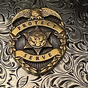 Puede incluir: Una hebilla de cintur&oacute;n de plata con un dise&ntilde;o ornamentado que presenta un escudo dorado con un &aacute;guila, una estrella y las palabras "PROTECT SERVE".
