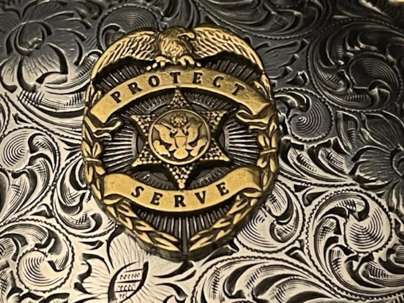 Puede incluir: Una hebilla de cintur&oacute;n de plata con un dise&ntilde;o ornamentado que presenta un escudo dorado con un &aacute;guila, una estrella y las palabras "PROTECT SERVE".