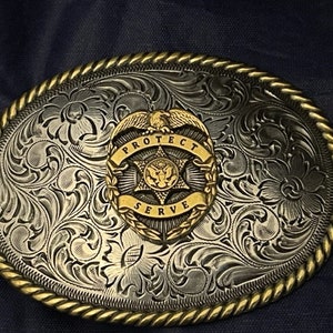 Puede incluir: Hebilla de cintur&oacute;n de plata con un dise&ntilde;o floral ornamentado y una insignia dorada que dice "Proteger y servir" con una estrella y un &aacute;guila.