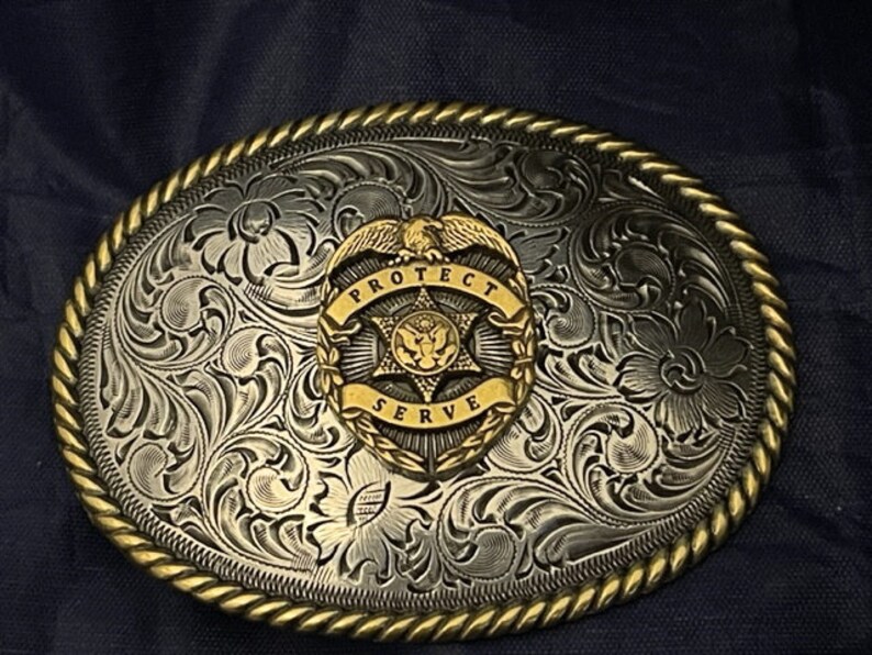 Puede incluir: Hebilla de cintur&oacute;n de plata con un dise&ntilde;o floral ornamentado y una insignia dorada que dice "Proteger y servir" con una estrella y un &aacute;guila.