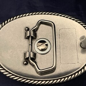 Puede incluir: Hebilla de cintur&oacute;n de plata con un borde de dise&ntilde;o de cuerda. La hebilla tiene un cierre de metal plateado y un &aacute;rea rectangular en blanco para grabar.