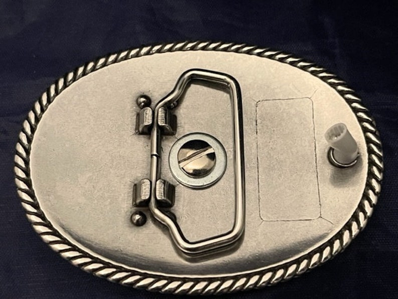 Puede incluir: Hebilla de cintur&oacute;n de plata con un borde de dise&ntilde;o de cuerda. La hebilla tiene un cierre de metal plateado y un &aacute;rea rectangular en blanco para grabar.