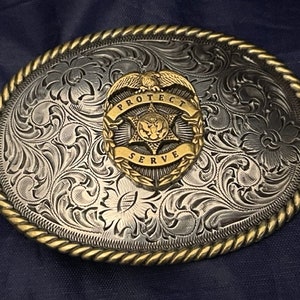 Puede incluir: Una hebilla de cintur&oacute;n de plata con un dise&ntilde;o ornamentado y un emblema dorado que dice "PROTECT SERVE".