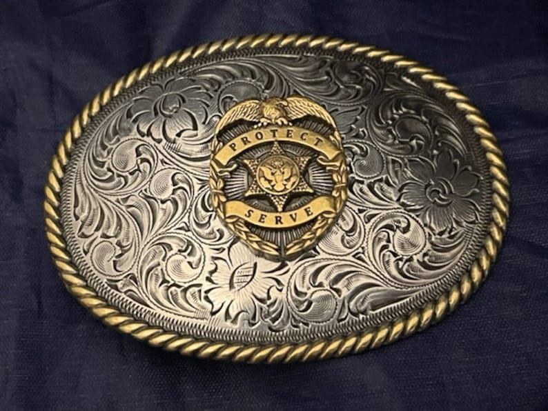 Puede incluir: Una hebilla de cintur&oacute;n de plata con un dise&ntilde;o ornamentado y un emblema dorado que dice "PROTECT SERVE".