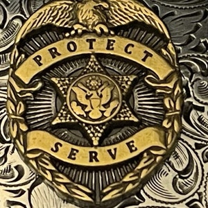 Puede incluir: Una insignia de metal dorado con una estrella de cinco puntas en el centro. La estrella tiene un dise&ntilde;o de &aacute;guila en su interior. La insignia tiene una pancarta que dice "PROTECT SERVE" alrededor de la estrella.