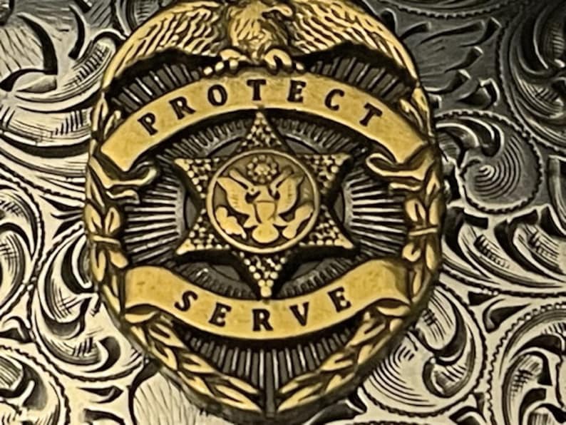 Puede incluir: Una insignia de metal dorado con una estrella de cinco puntas en el centro. La estrella tiene un dise&ntilde;o de &aacute;guila en su interior. La insignia tiene una pancarta que dice "PROTECT SERVE" alrededor de la estrella.
