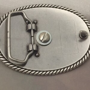May include: Silver belt buckle with a rope-like border and a metal loop on the back. The buckle has a blank space for engraving.