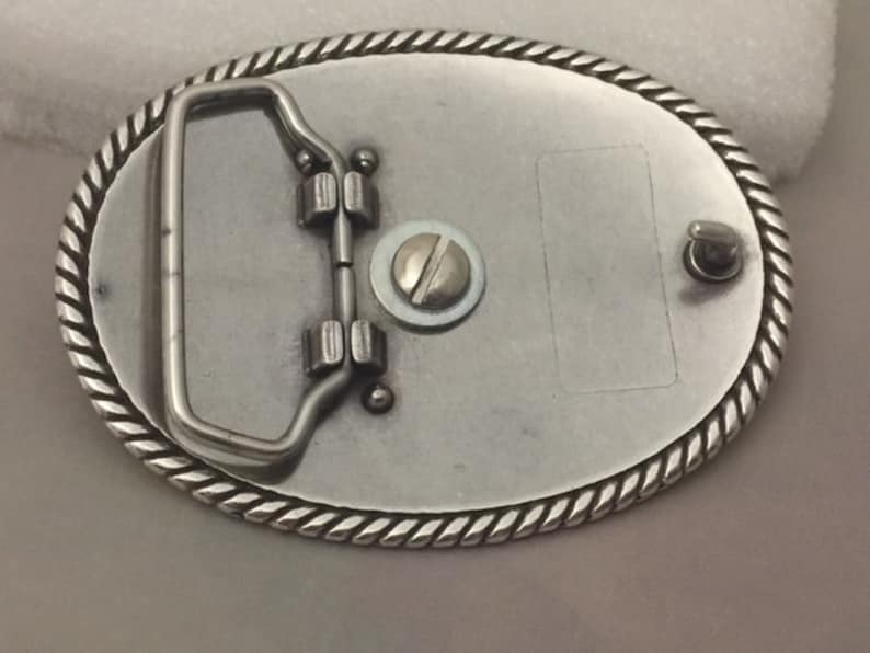 May include: Silver belt buckle with a rope-like border and a metal loop on the back. The buckle has a blank space for engraving.