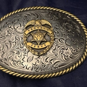 Puede incluir: Hebilla de cintur&oacute;n de plata con un dise&ntilde;o grabado en tono dorado. La hebilla presenta una insignia en forma de estrella con las palabras "Protect and Serve" alrededor del borde.