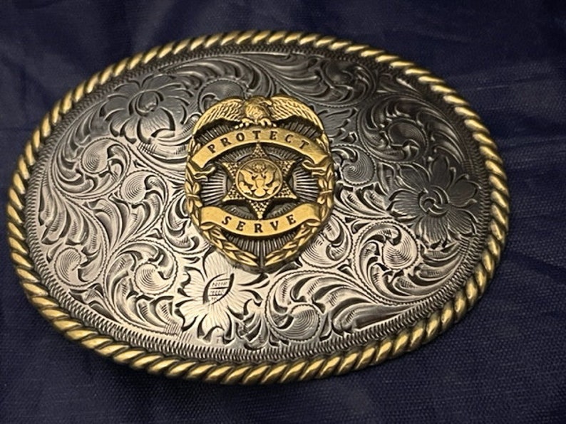 Puede incluir: Hebilla de cintur&oacute;n de plata con un dise&ntilde;o grabado en tono dorado. La hebilla presenta una insignia en forma de estrella con las palabras "Protect and Serve" alrededor del borde.