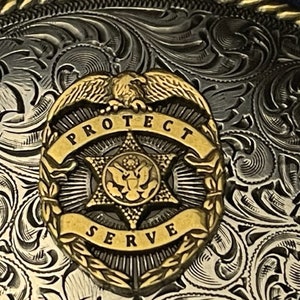 Puede incluir: Una hebilla de cintur&oacute;n de plata y oro con un intrincado dise&ntilde;o floral. El centro de la hebilla presenta una estrella dorada con un escudo y las palabras "Proteger y Servir".