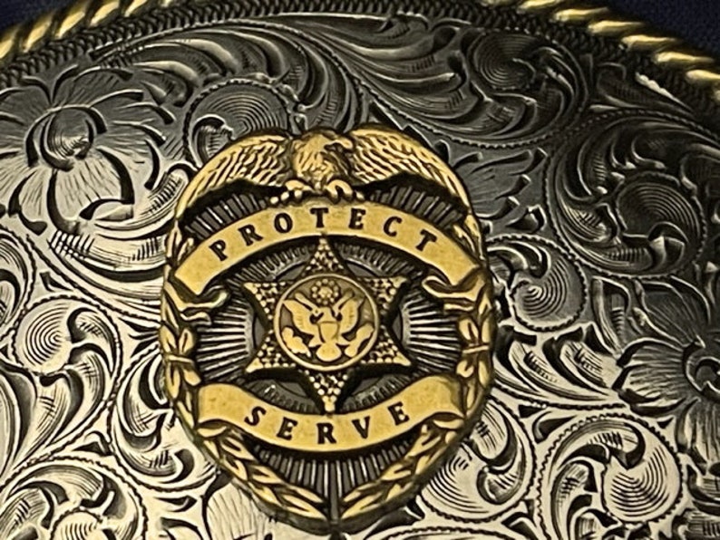 Puede incluir: Una hebilla de cintur&oacute;n de plata y oro con un intrincado dise&ntilde;o floral. El centro de la hebilla presenta una estrella dorada con un escudo y las palabras "Proteger y Servir".